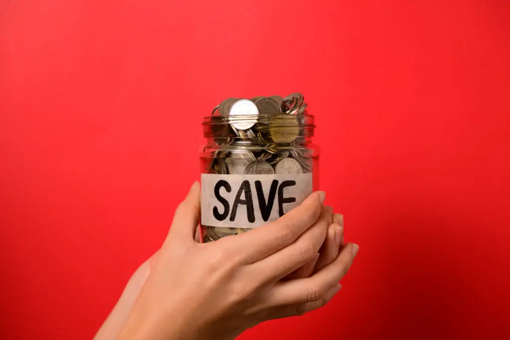 Individual holding up a glass jar filled with coins that bears a sign with the word "save".