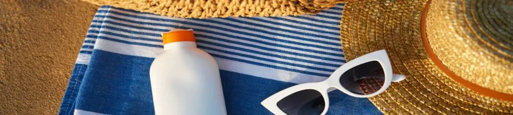 A bottle of sunscreen and sunglasses lying on top of a towel on a sandy beach.