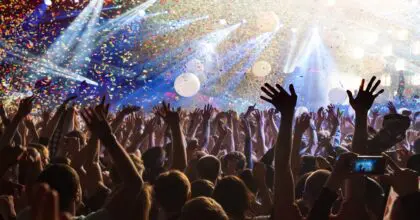 A busy crowd at a concert with their hands up in the air, spotlights from the stage at the front shining on them and confetti raining down on them.