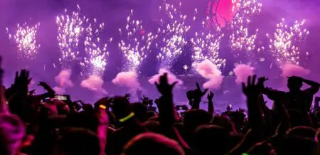 Concert crowd with their arms up, purple lights illuminating the scene and fireworks going off in front of the stage.