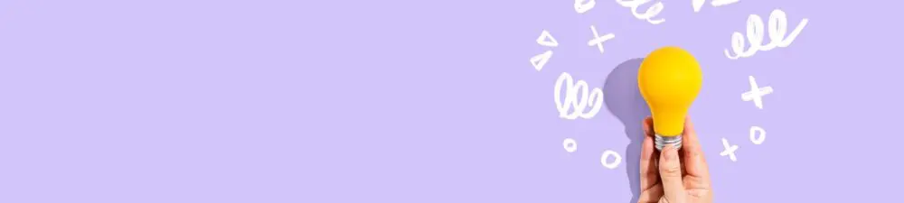 A hand holds a lit, yellow lightbulb against a purple background to signify innovation.