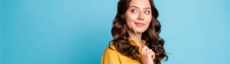 Woman wearing a yellow blouse, standing in front of blue background, holding a pencil & looking thoughtful
