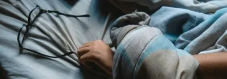 Halb zugedeckter Arm auf einer Matratze. Brille liegt links neben der Hand.