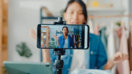 Young Asia lady fashion designer using mobile phone receiving purchase order and showing clothes recording video live streaming online at shop.