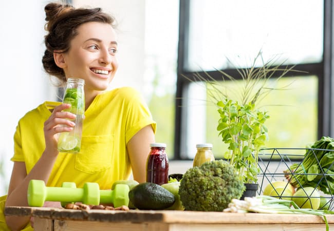 Woman drinking a functional drink while sitting down surrounded by vegetables. Source: GettyImages