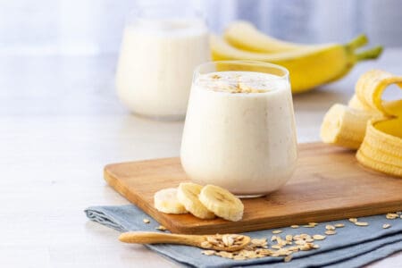 A glass of banana and oat yoghurt smoothie on a wooden board, surrounded by the ingredients.