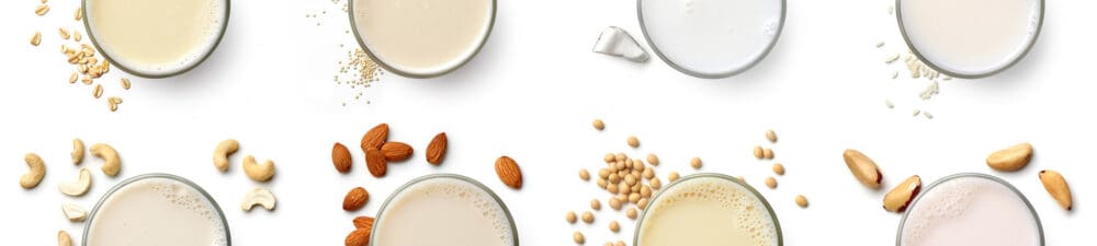 Glasses of plant based milks with assorted nuts and grains surrounding them.