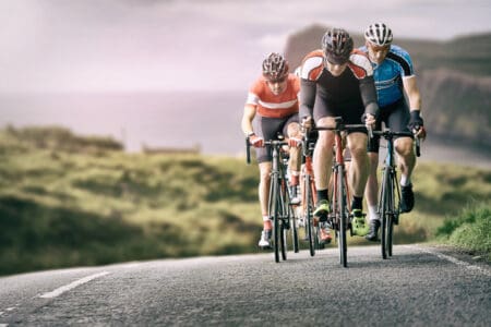 A group of cyclists ride their bikes along a coastal road with grassy cliffs in the background.