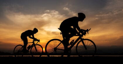 Two people silhouetted against a sunset, riding bicycles and wearing cycling helmets.