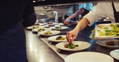 Chefs plate up dishes of fish and greens on a service area of a restaurant kitchen.