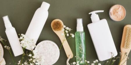 Assortment of bottles containing personal care products alongside brushes on a green background, framed by white flowers.