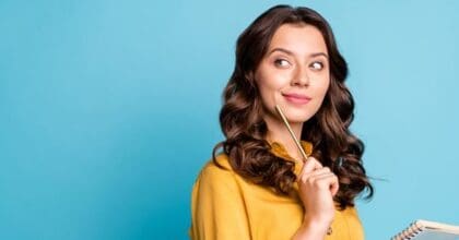 Woman wearing a yellow blouse, standing in front of blue background, holding a pencil & looking thoughtful